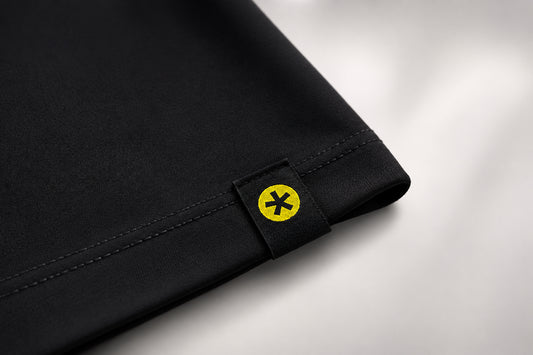 Close-up of a black fabric product corner with a small black tab featuring a yellow circle and black asterisk symbol, set against a light gray background.